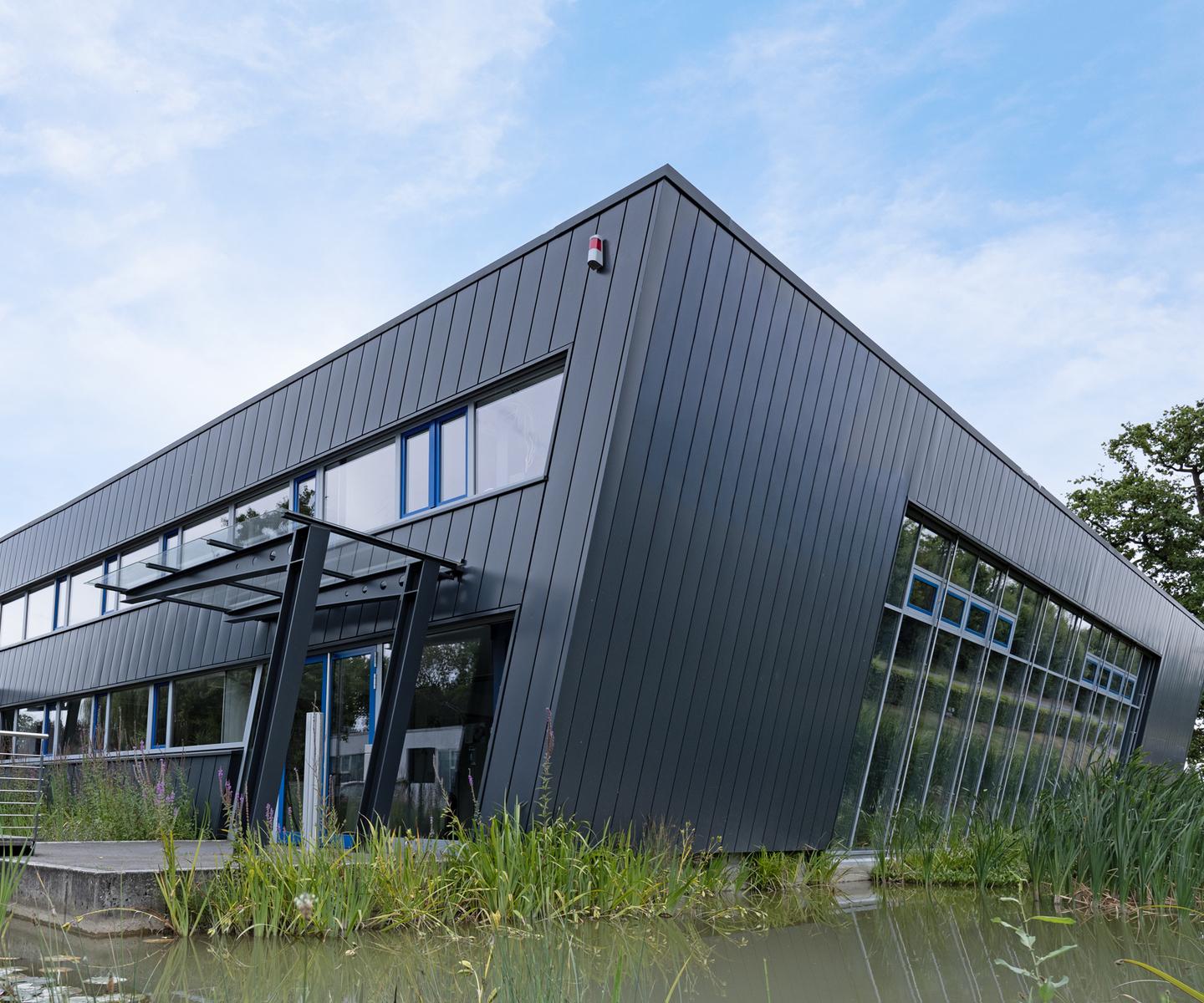 Moderne, schwarze Geb&auml;udeansicht mit gro&szlig;en Fenstern, umgeben von grasbewachsenen Uferbereichen und Wasserpflanzen.