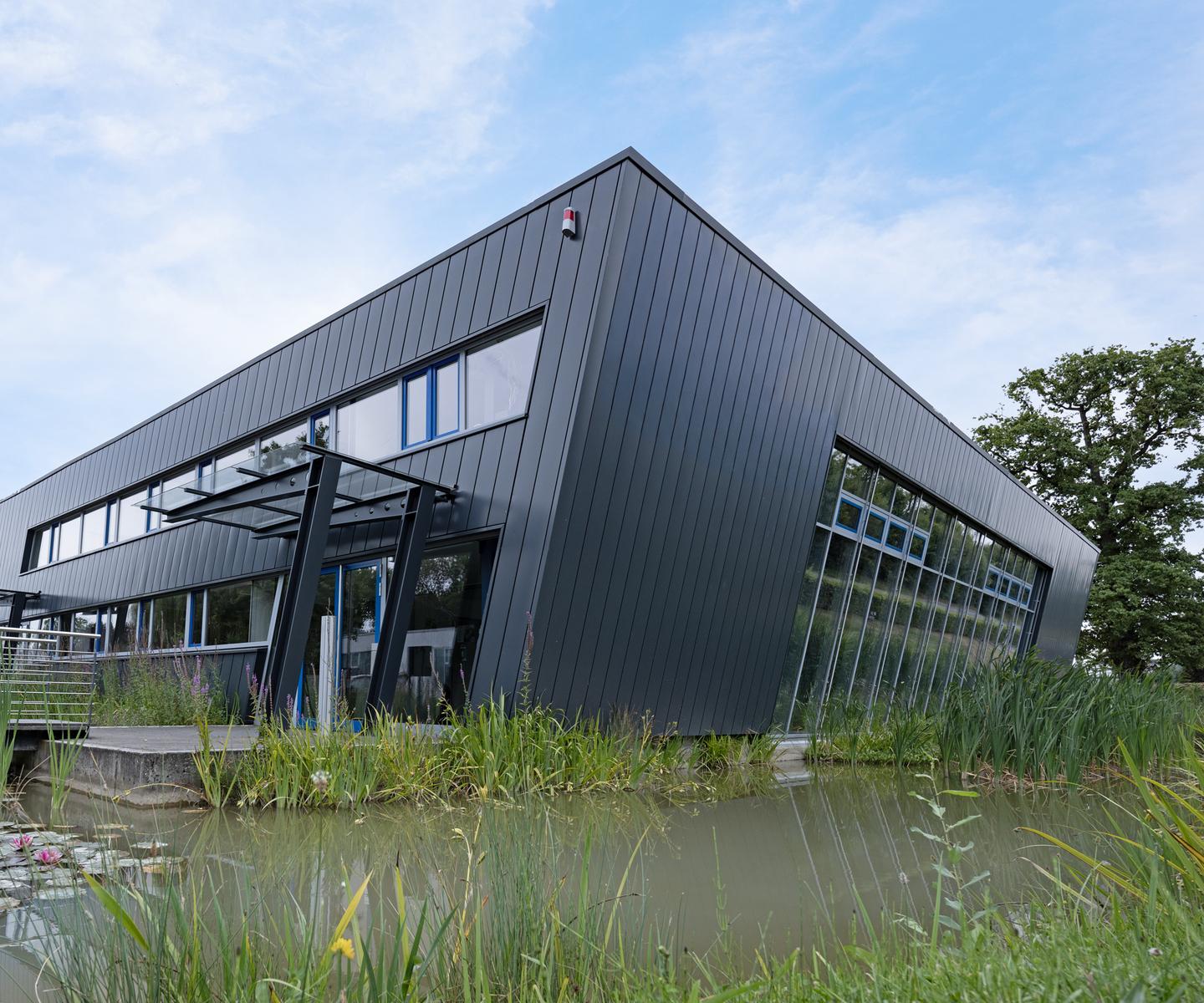 Moderne, schwarze Gebäudeansicht mit großen Fenstern, umgeben von grasbewachsenen Uferbereichen und Wasserpflanzen.