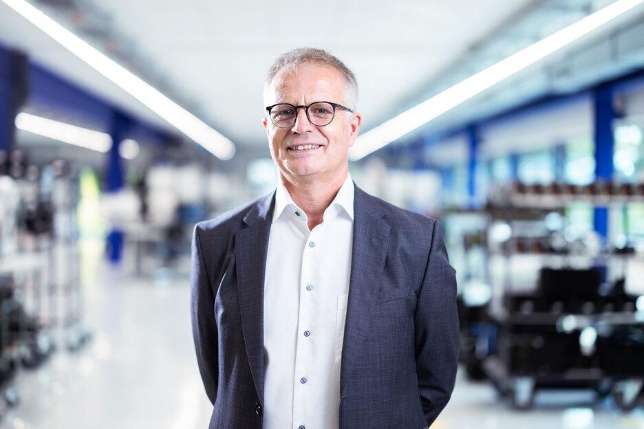 Portrait eines Mannes in Anzug mit Brille, lächeln, im Hintergrund ein moderner, heller Arbeitsplatz.