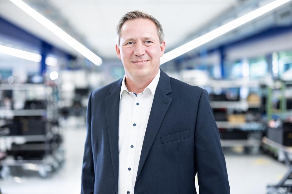 Portrait eines Mannes in einem dunklen Anzug vor unscharfer Werkstattmitte mit Regalen im Hintergrund.