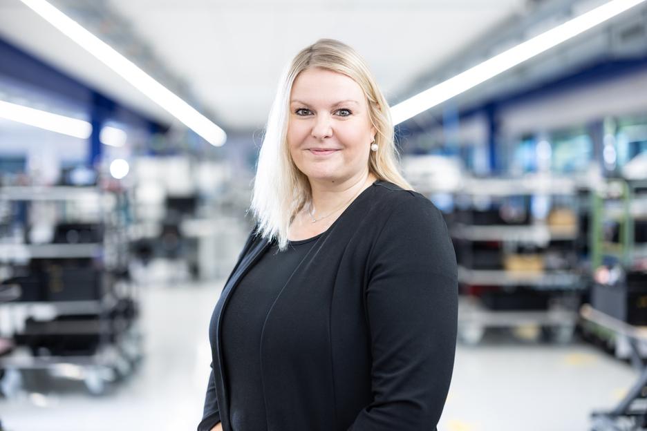 Portr&auml;t einer l&auml;chelnden Frau mit blonden Haaren in einem modernen, industriellen Umfeld mit Regalen im Hintergrund.