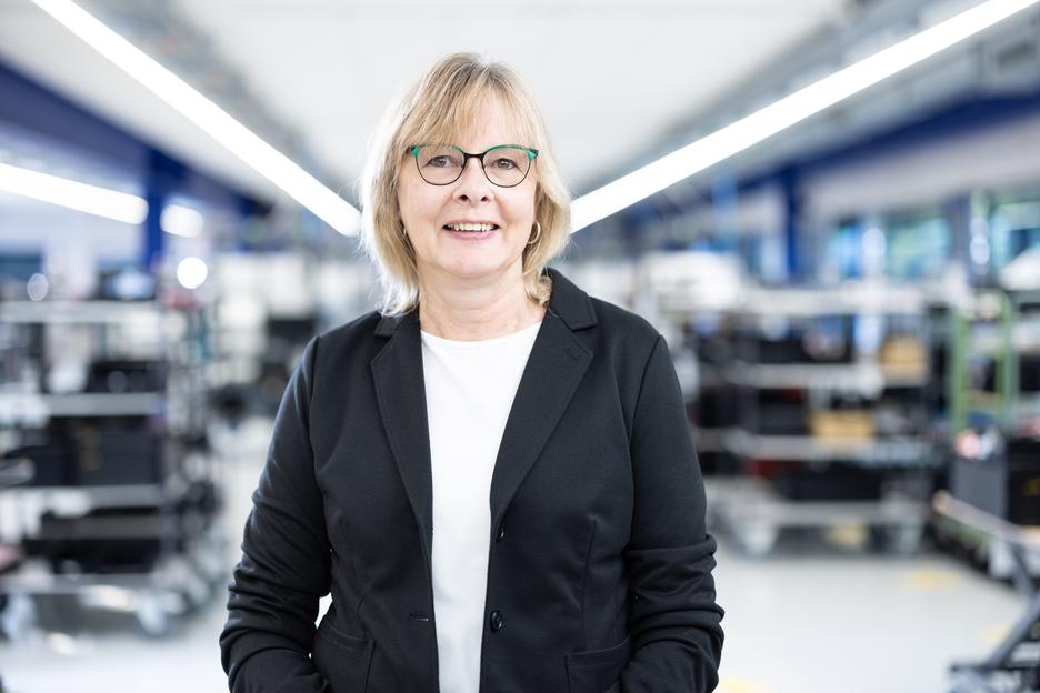 Portr&auml;t einer l&auml;chelnden Frau mit Brille, in schwarzer Jacke, vor industriellem Hintergrund mit Regalen.