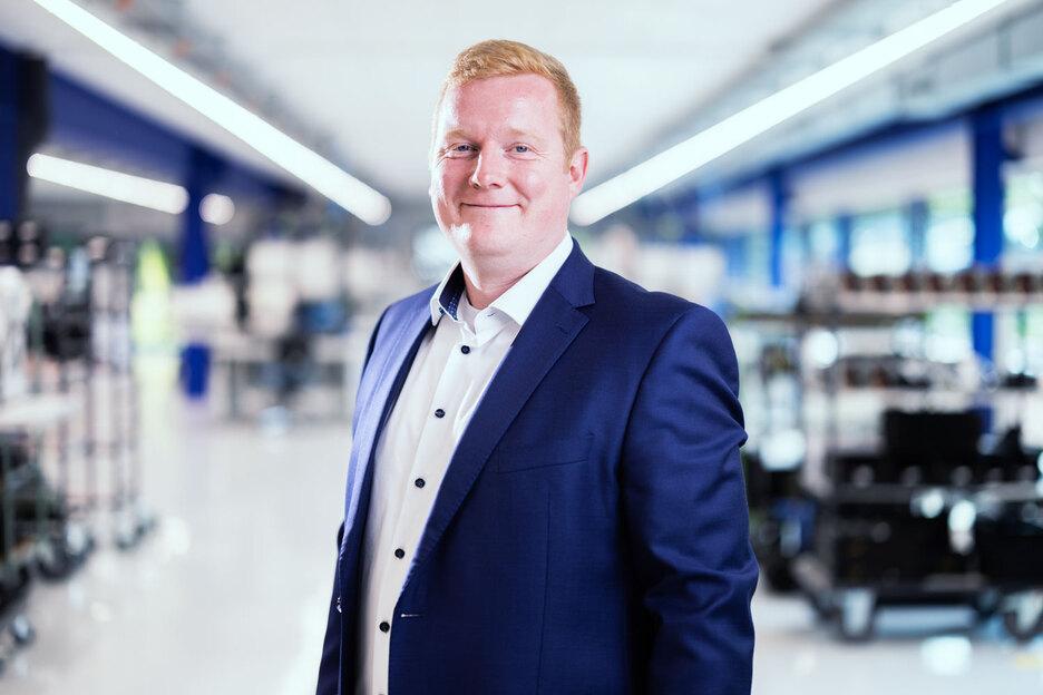 Mann in Anzug l&auml;chelt in einem modernen, hellen Raum mit Regalen im Hintergrund.