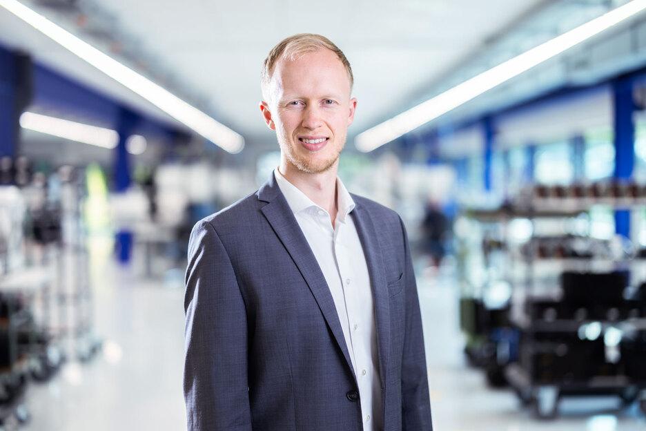 Portr&auml;t eines Mannes in einem grauen Anzug, vor einem modernen, hellen Hintergrund mit Regalen und Arbeitsbereichen.