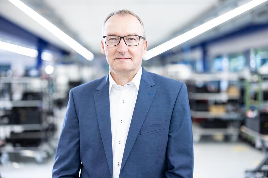 Portr&auml;t eines Mannes in einem blauen Anzug vor einem industriellen Hintergrund mit Regalen und ged&auml;mpften Lichtern.