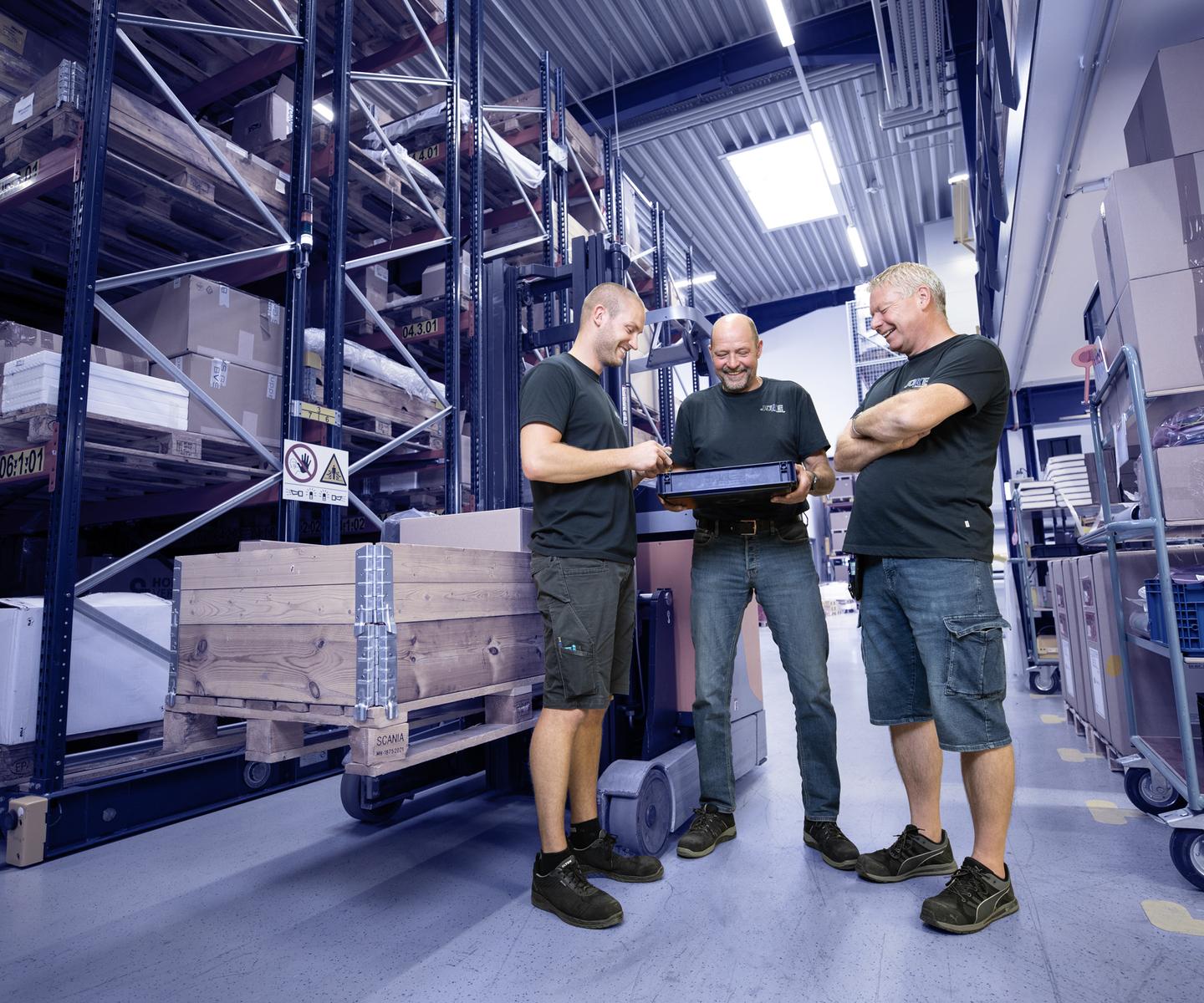 Drei Männer in Arbeitskleidung schauen auf einen Laptop in einem Lager mit Regalen voller Kisten und Paletten.