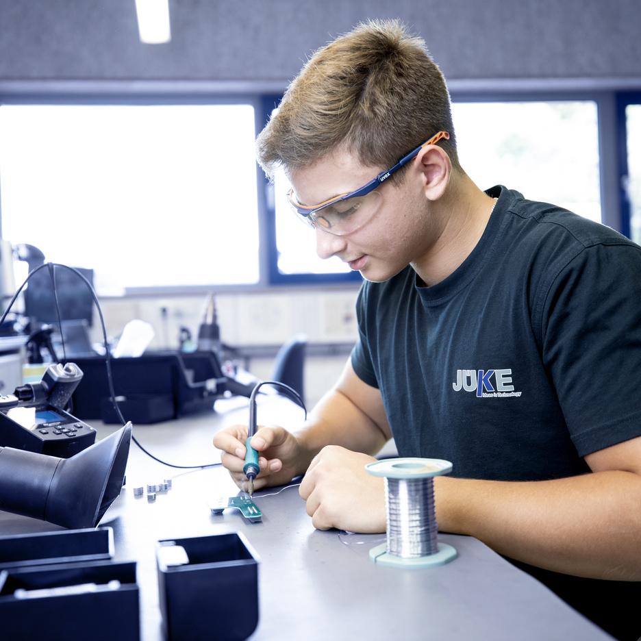 Junger Mann mit Schutzbrille l&ouml;tet an einer Leiterplatte in einer Werkstatt, mit Kabeln und Werkzeugen auf dem Tisch.