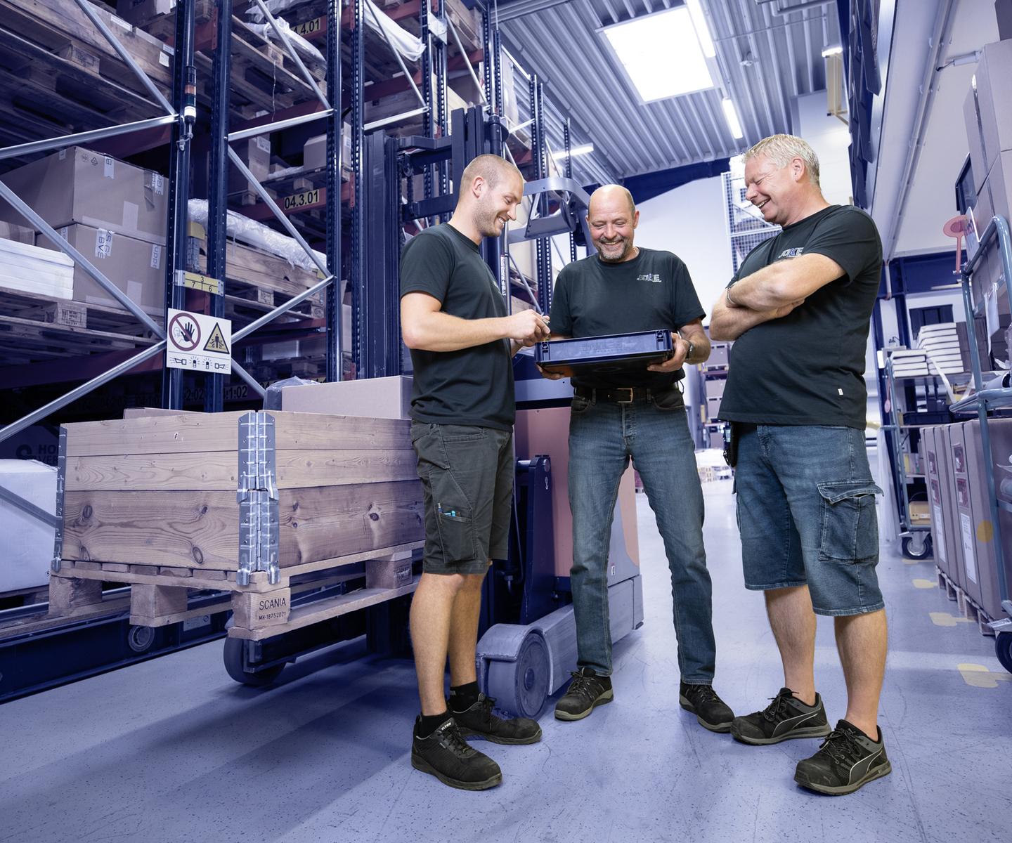 Drei M&auml;nner in Arbeitskleidung schauen auf einen Laptop in einem Lager mit Regalen voller Kisten und Paletten.