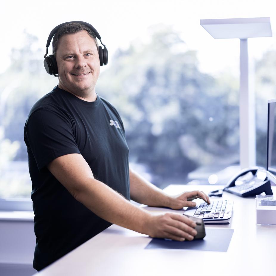 Mann in schwarzem T-Shirt mit Kopfhörern steht an einem Schreibtisch und bedient eine Computermaus und -tastatur.