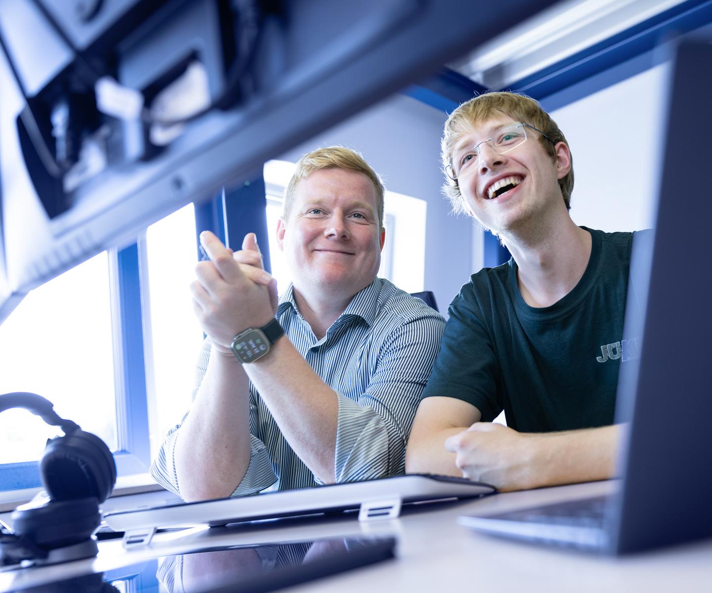 Zwei Männer lächeln und schauen auf einen Computerbildschirm, während sie an einem Tisch mit Laptop und Kopfhörern sitzen.