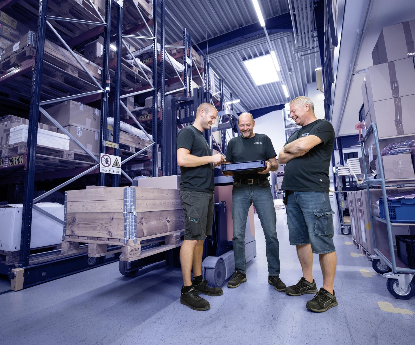 Three men in workwear look at a laptop in a warehouse with shelves full of boxes and pallets.