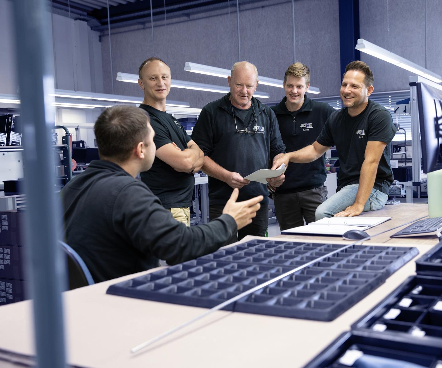 Gruppe von f&uuml;nf M&auml;nnern in Arbeitskleidung diskutiert um einen Tisch mit Dokumenten und Werkzeugen in einer Werkstatt.