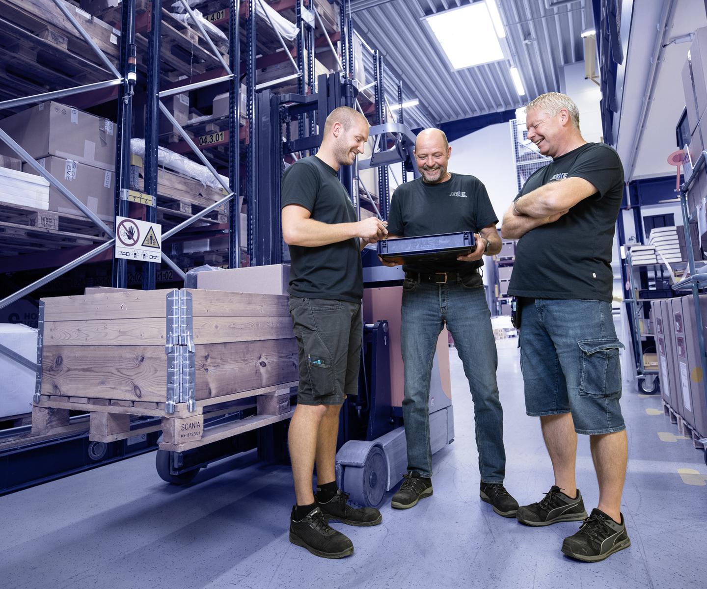 Drei M&auml;nner in Arbeitskleidung schauen auf einen Laptop in einem Lager mit Regalen voller Kisten und Paletten.