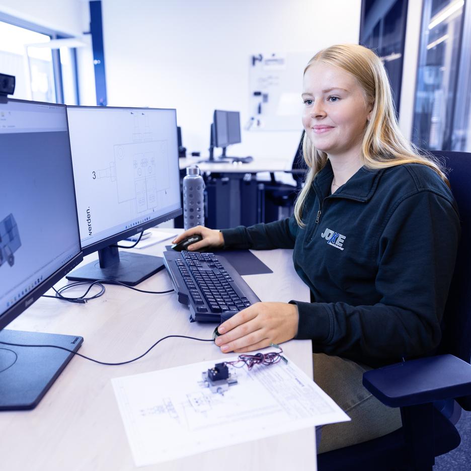 Junge Frau sitzt an einem Tisch mit zwei Monitoren, arbeitet an CAD-Software und hat technische Zeichnungen vor sich.