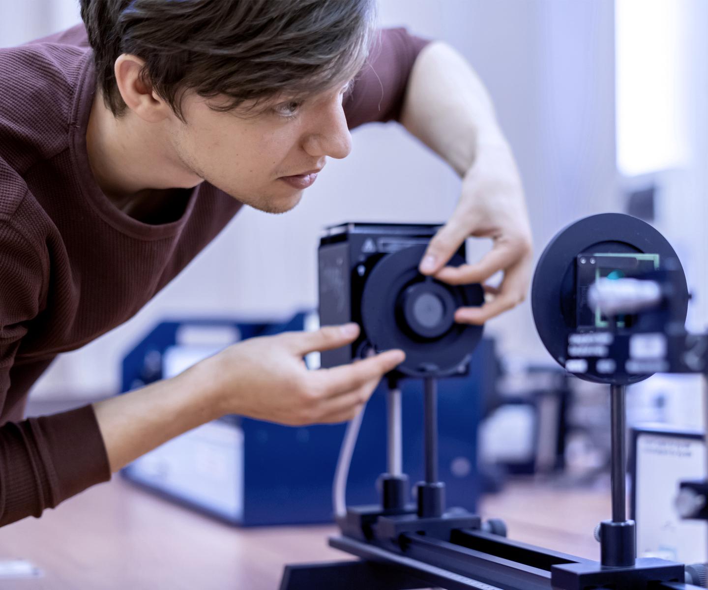 Junger Mann justiert ein optisches Ger&auml;t auf einem Tisch in einem Laborumfeld.