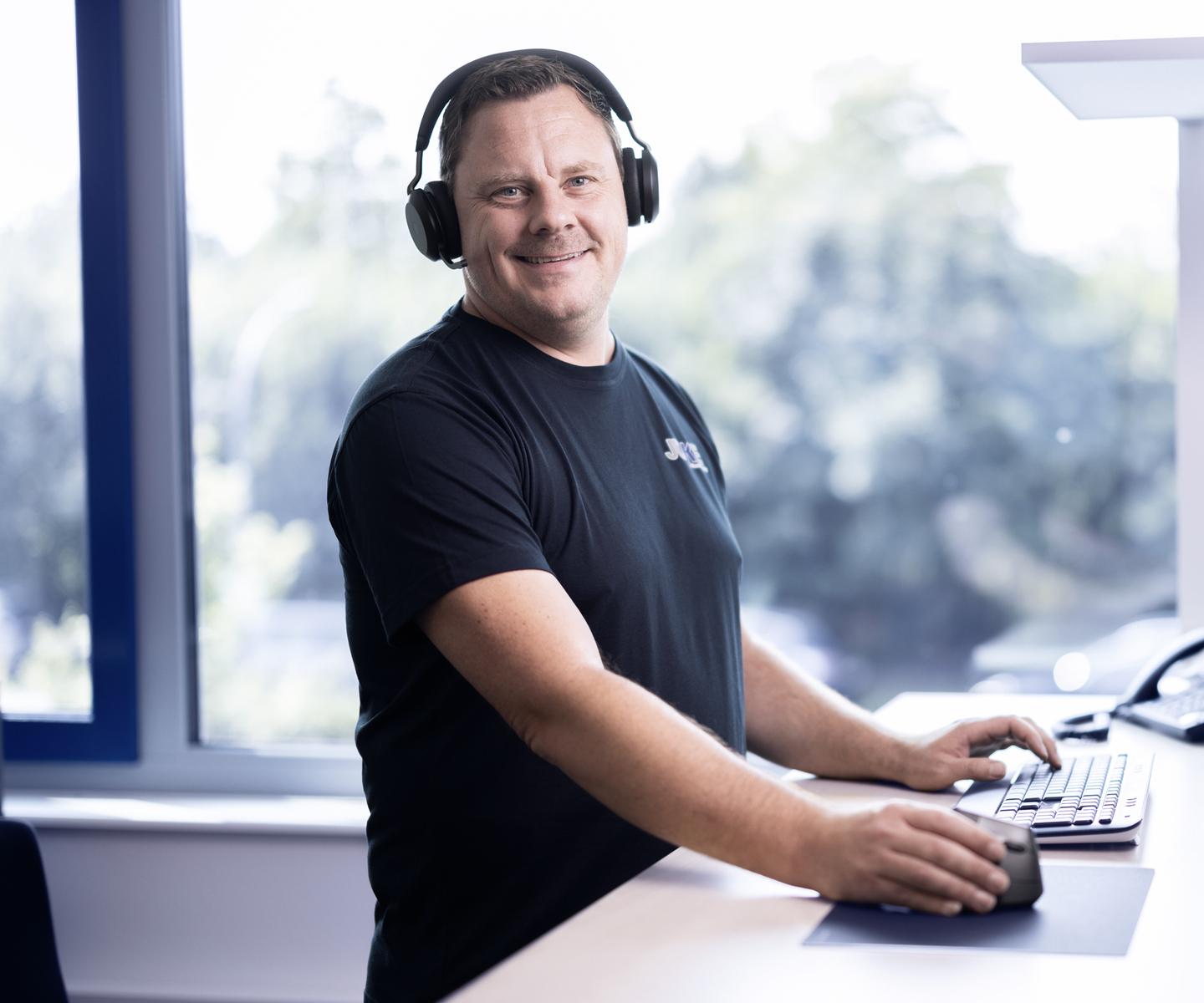 Mann mit Kopfh&ouml;rern arbeitet an einem Schreibtisch mit Computern und Telefon in einem hellen B&uuml;ro.