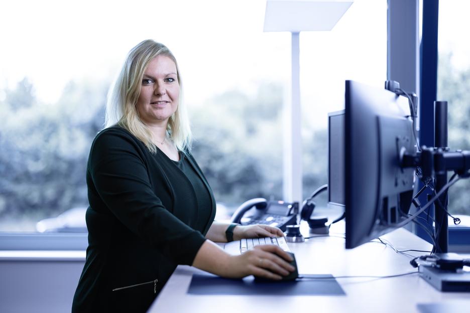 Frau mit blonden Haaren arbeitet an einem Schreibtisch mit Computer und Telefon in einem hellen B&uuml;ro.