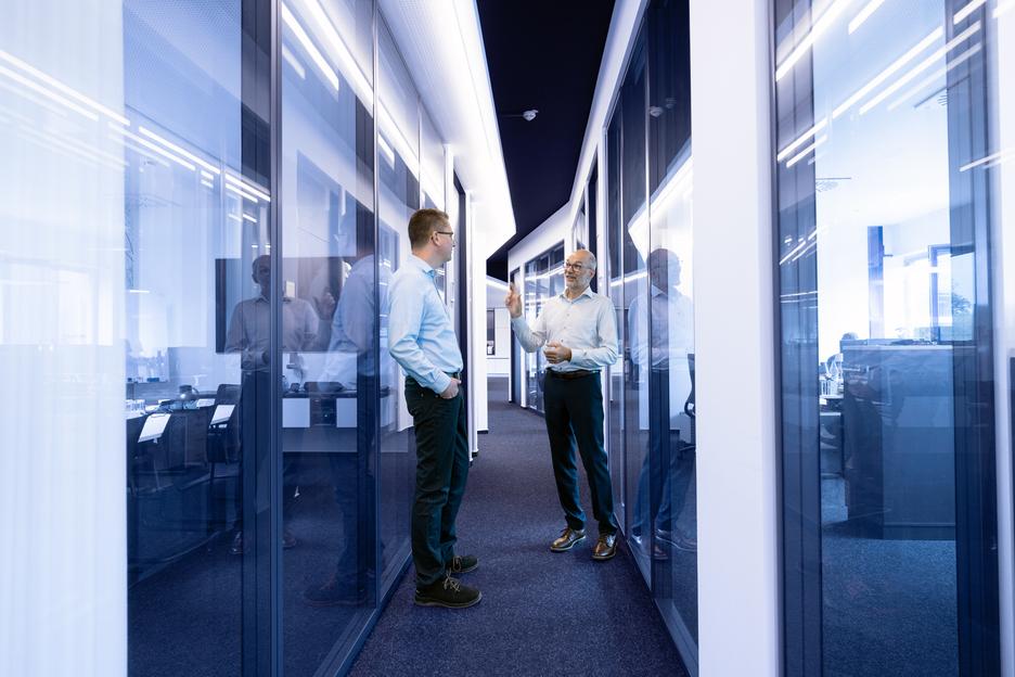 Zwei Männer in Bürokleidung stehen in einem modernen Flur mit Glasseitenwänden und diskutieren miteinander.
