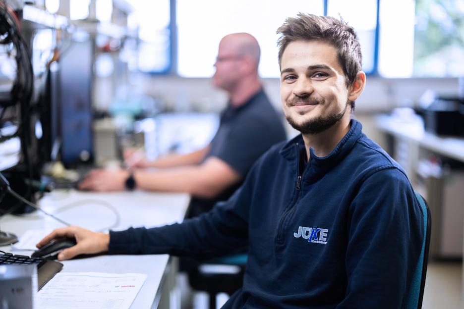 Lächelnder Mann im blauen Pullover sitzt an einem Tisch mit Computer, im Hintergrund ein weiterer Kollege bei der Arbeit.