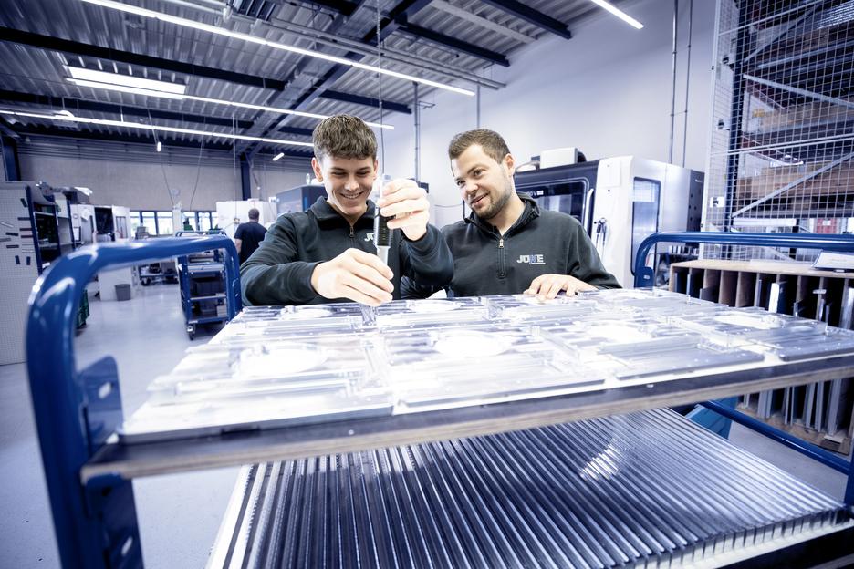 Zwei Männer überprüfen gemeinsam transparente Teile auf einem Werkstattwagen in einer modernen Fertigungsumgebung.