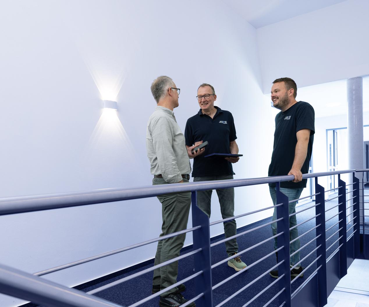 Drei M&auml;nner stehen auf einer Treppe und unterhalten sich, ein Tablet in der Hand, im modernen, hellen Raum.