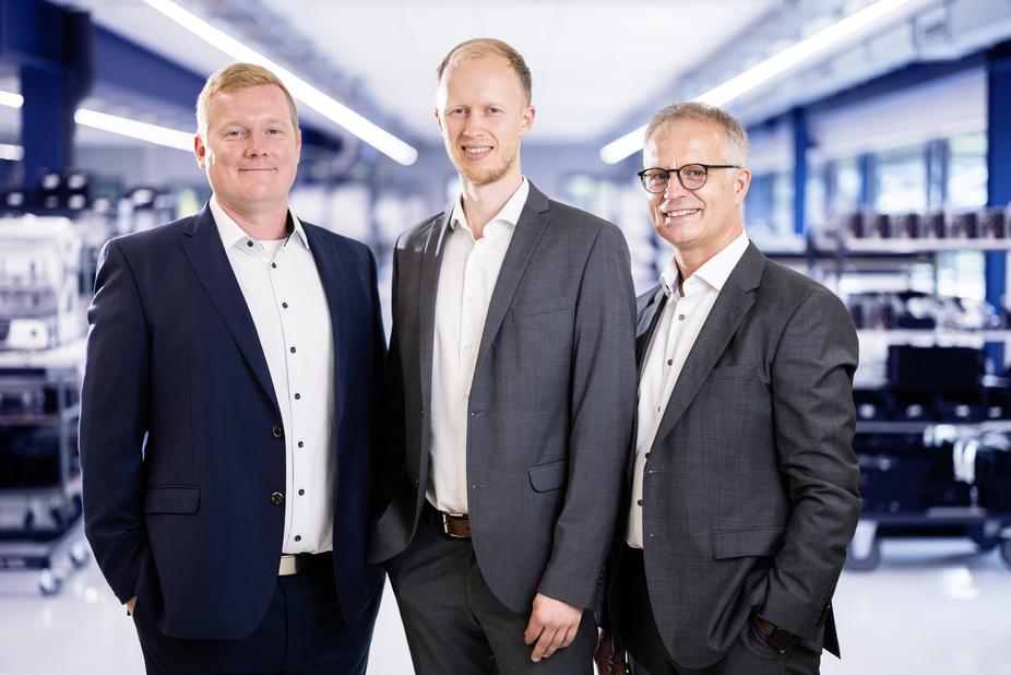 Drei M&auml;nner in Anz&uuml;gen stehen in einem modernen B&uuml;ro mit technischen Ger&auml;ten im Hintergrund.