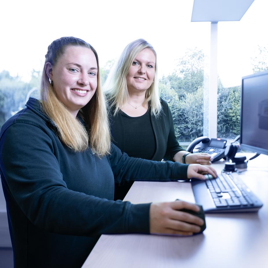 Zwei Frauen sitzen an einem Schreibtisch, arbeiten am Computer und l&auml;cheln in die Kamera. Hintergrund mit B&auml;umen.