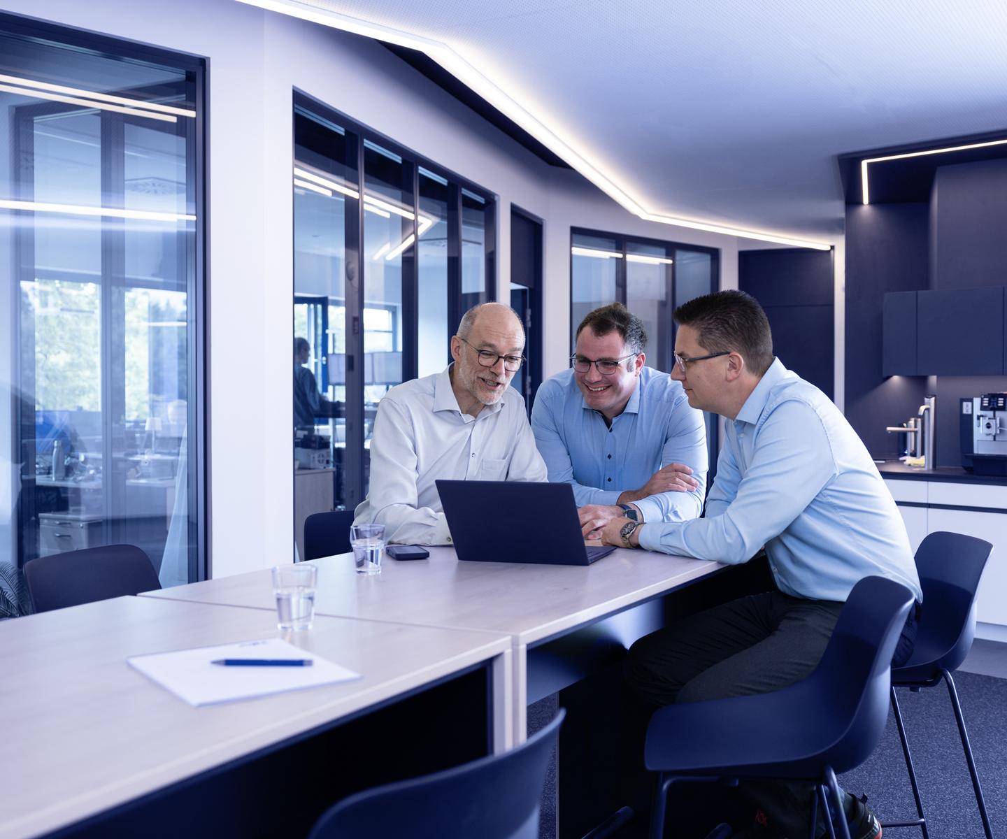 Drei M&auml;nner in Hemden sitzen an einem Tisch und betrachten gemeinsam einen Laptop im modernen B&uuml;ro.