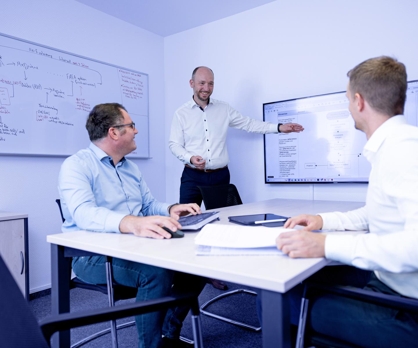 Drei Personen in einem Meetingraum diskutieren, w&auml;hrend einer an einem Bildschirm eine Pr&auml;sentation zeigt.