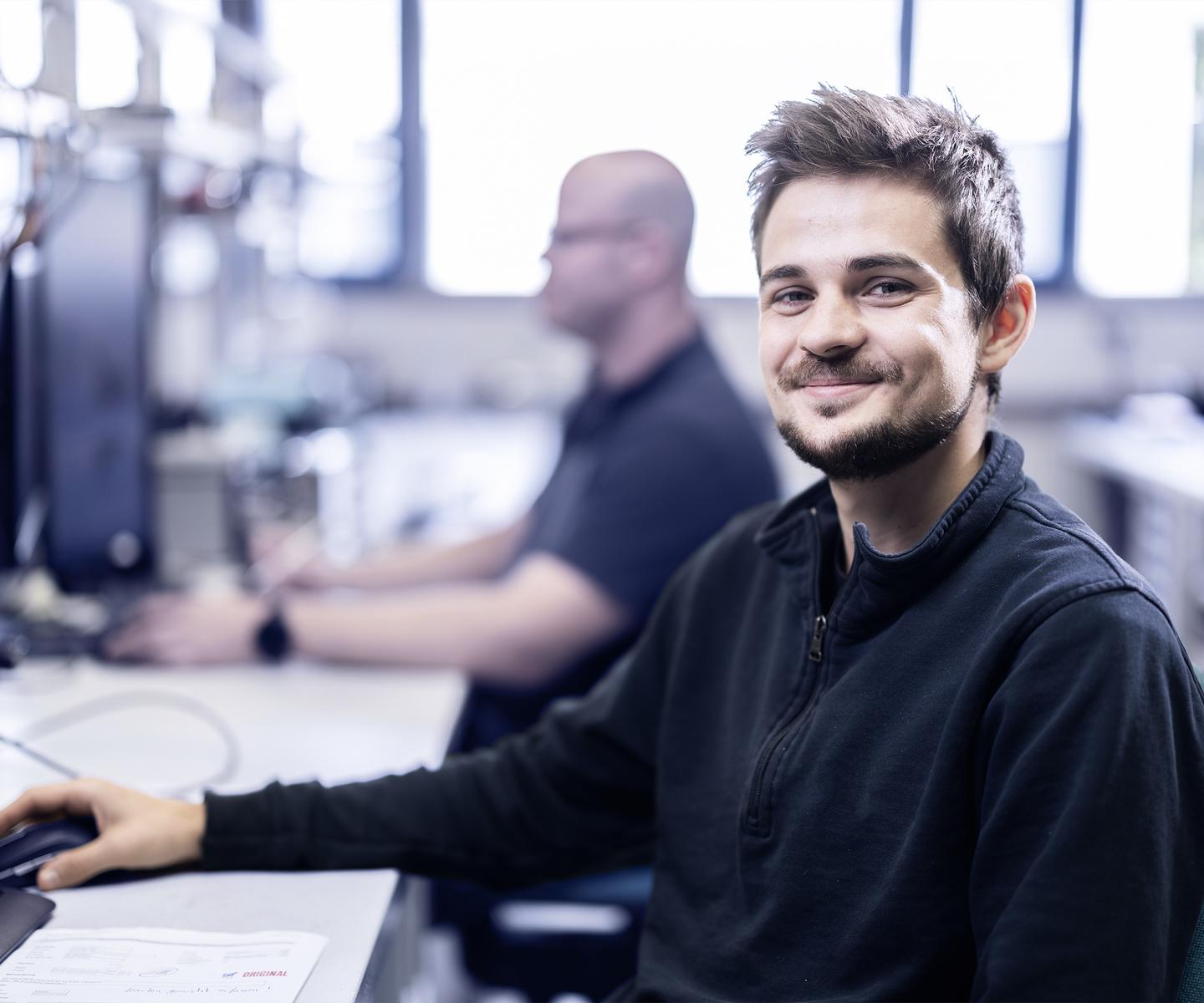 L&auml;chelnder junger Mann sitzt an einem Schreibtisch, vor ihm ein Computer, im Hintergrund ein weiterer Kollege.