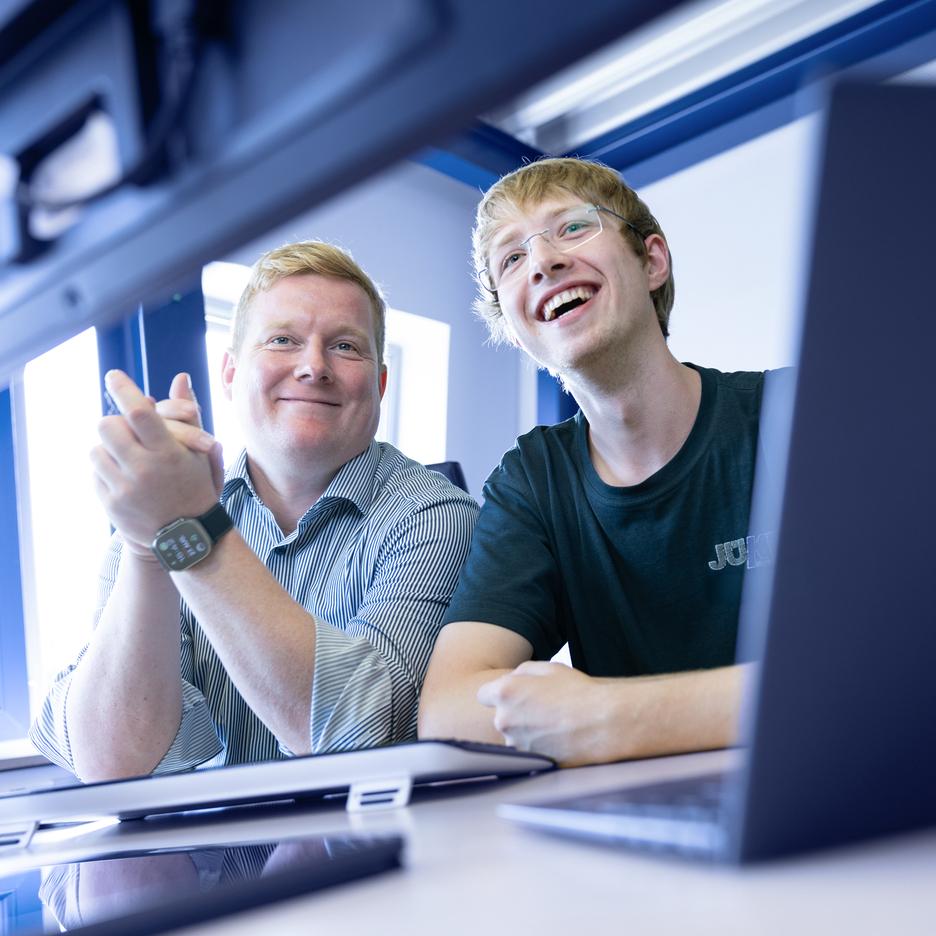 Zwei Männer lächeln und schauen auf einen Computerbildschirm, während sie an einem Tisch mit Laptop und Kopfhörern sitzen.