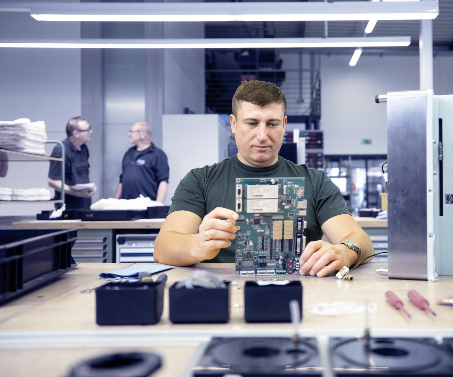 Man in a T-shirt examining a circuit board at a table while two colleagues talk in the background.