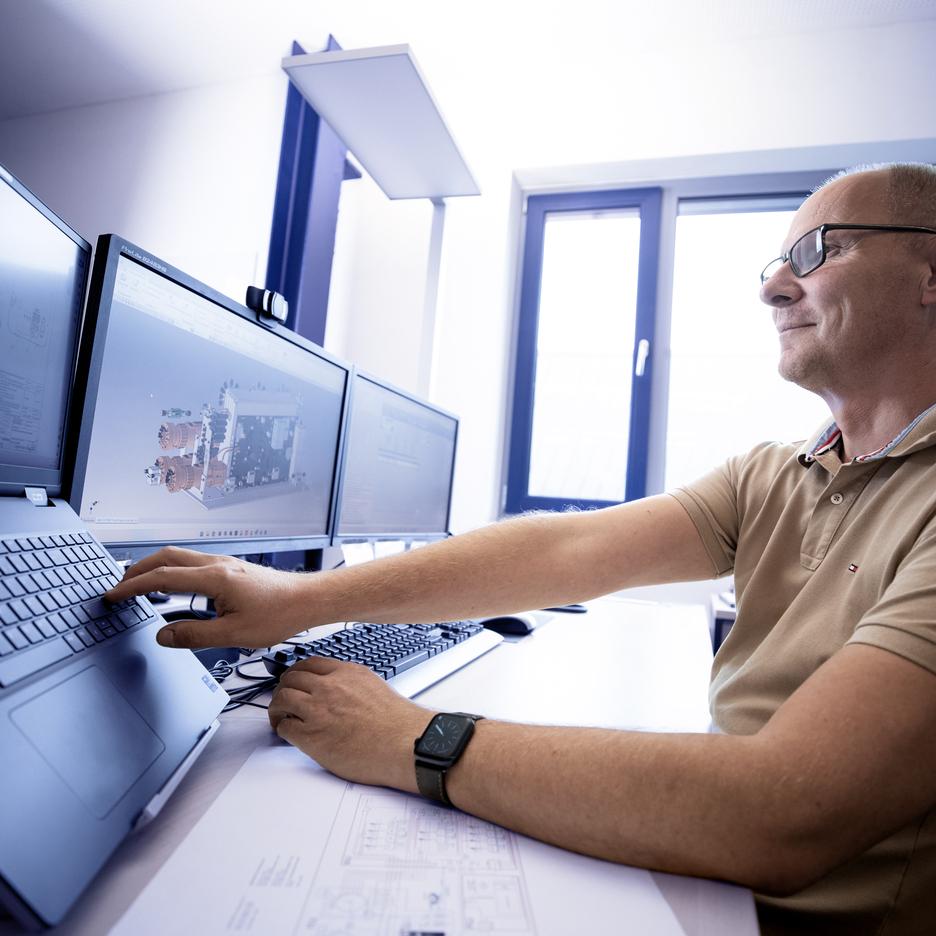 Mann mit Brille arbeitet an mehreren Monitoren, die technische Zeichnungen und Modelle zeigen.