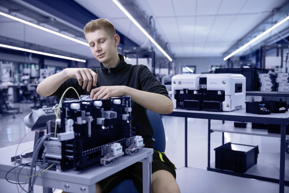 Junger Mann arbeitet an einem technischen Ger&auml;t in einem modernen Labor mit medizinischen Ger&auml;ten im Hintergrund.