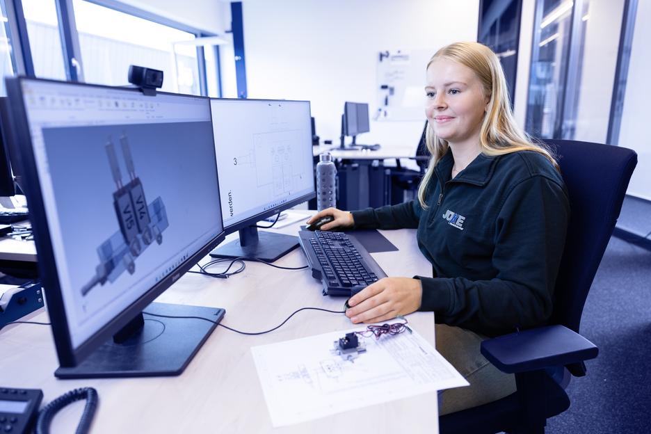 Junge Frau sitzt an einem Tisch mit zwei Monitoren, arbeitet an CAD-Software und hat technische Zeichnungen vor sich.