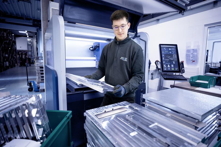 Junger Mann in Arbeitskleidung h&auml;lt bearbeitete Metallplatten vor einer Maschine in einer Produktionshalle.