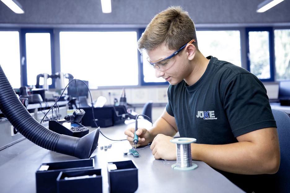 Junger Mann mit Schutzbrille l&ouml;tet an einer Leiterplatte in einer Werkstatt, mit Kabeln und Werkzeugen auf dem Tisch.