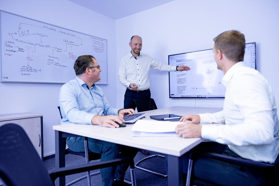 Drei Personen in einem Meetingraum diskutieren, w&auml;hrend einer an einem Bildschirm eine Pr&auml;sentation zeigt.