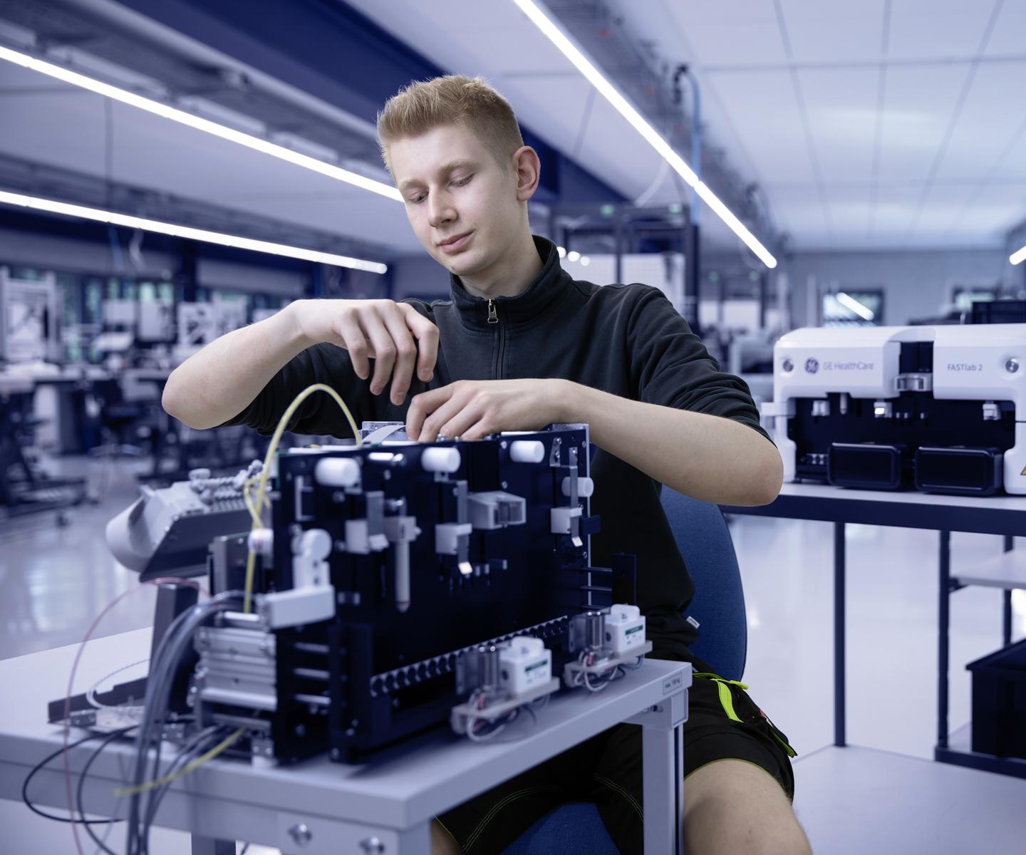 Junger Mann arbeitet an einem technischen Ger&auml;t in einem modernen Labor mit medizinischen Ger&auml;ten im Hintergrund.