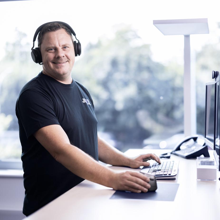 Mann mit Kopfh&ouml;rern arbeitet an einem Schreibtisch mit Computern und Telefon in einem hellen B&uuml;ro.