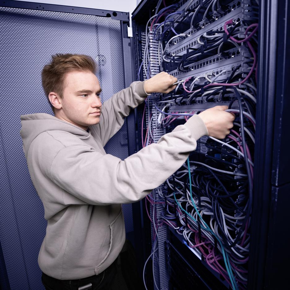 Junger Mann in grauem Hoodie verkabelt ein Server-Rack mit bunten Kabeln.