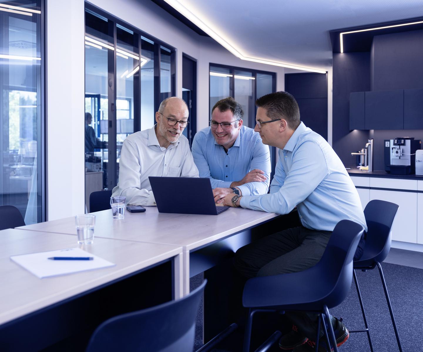 Drei M&auml;nner in Hemden sitzen an einem Tisch und betrachten gemeinsam einen Laptop im modernen B&uuml;ro.