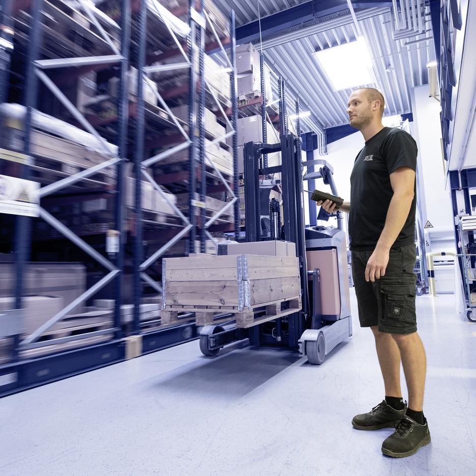 Mitarbeiter steht mit Handscanner vor einem Hochregal voller Kartons und einer Gabelstaplerpalette in einem Lager.