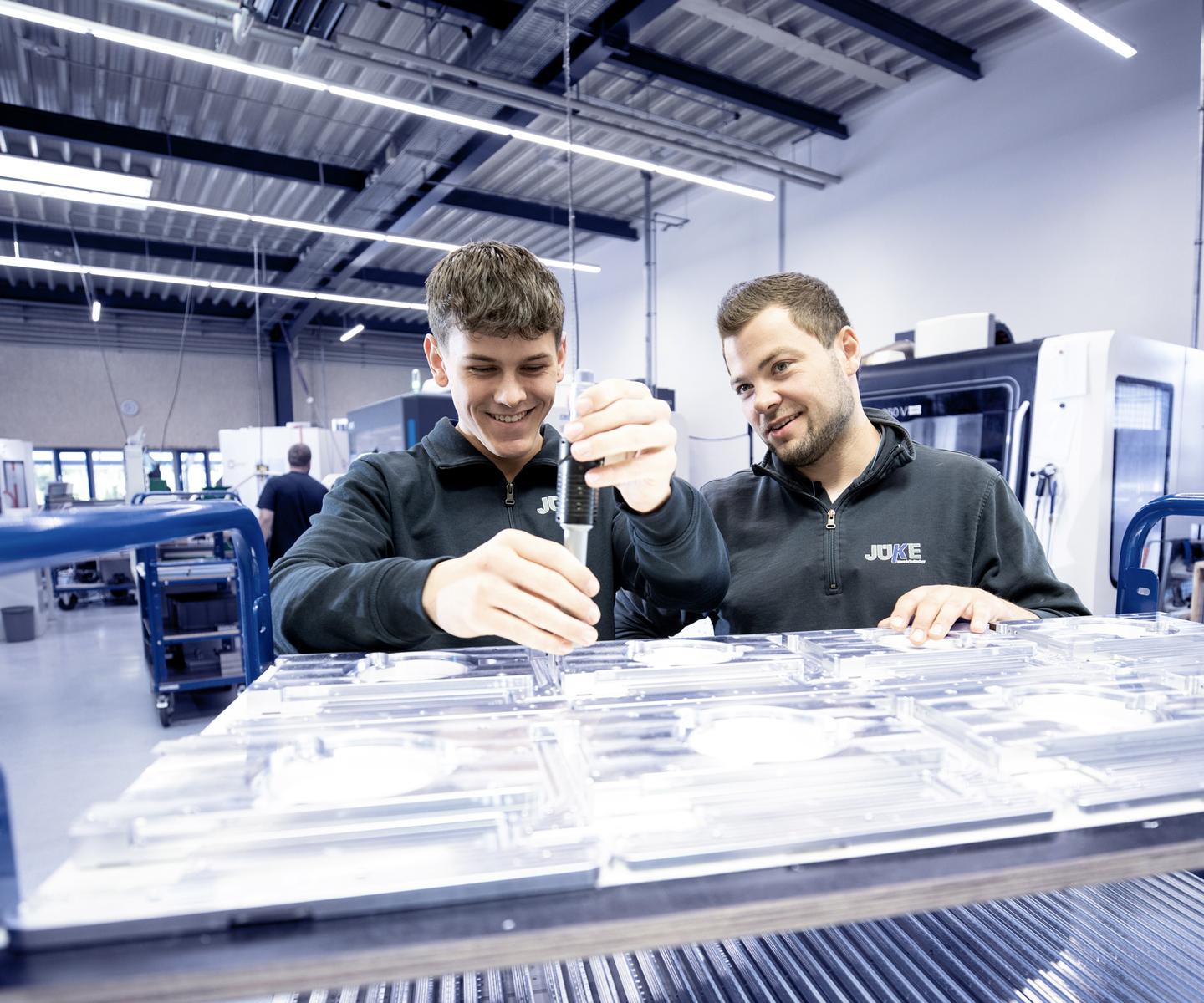 Zwei M&auml;nner &uuml;berpr&uuml;fen gemeinsam transparente Teile auf einem Werkstattwagen in einer modernen Fertigungsumgebung.