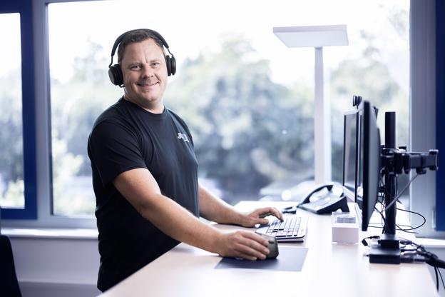 Mann mit Kopfh&ouml;rern arbeitet an einem Schreibtisch mit Computern und Telefon in einem hellen B&uuml;ro.