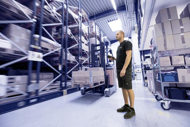 Mitarbeiter steht mit Handscanner vor einem Hochregal voller Kartons und einer Gabelstaplerpalette in einem Lager.