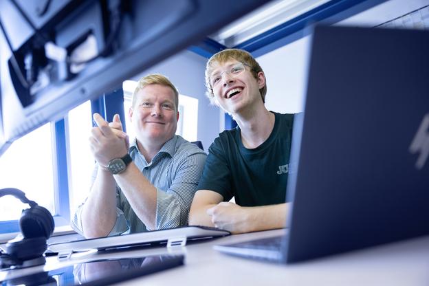 Zwei Männer lächeln und schauen auf einen Computerbildschirm, während sie an einem Tisch mit Laptop und Kopfhörern sitzen.