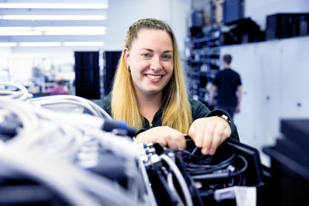 Frau mit langen, blonden Haaren arbeitet l&auml;chelnd an einer Verkabelung in einem modernen technischen Raum.