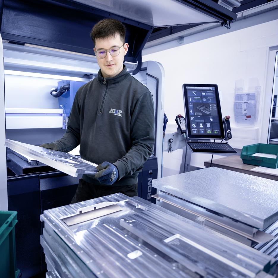 Junger Mann in Arbeitskleidung hält bearbeitete Metallplatten vor einer Maschine in einer Produktionshalle.