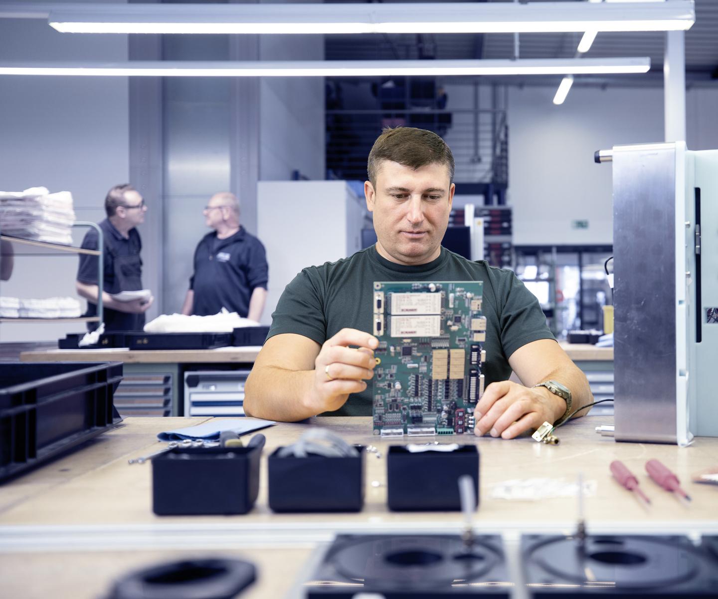 Mann in T-Shirt betrachtet eine Platine an einem Tisch, w&auml;hrend zwei Kollegen im Hintergrund miteinander sprechen.
